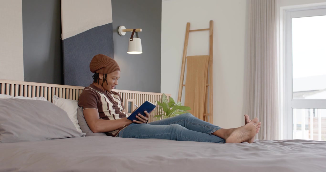 Adult African man holding blue paperback book, reading on king bed, shifting pages glancing window