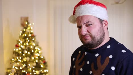 frowning young man with santa hat and a xmas sweater standing in front of a christmas tree with a disappointed expression on his face