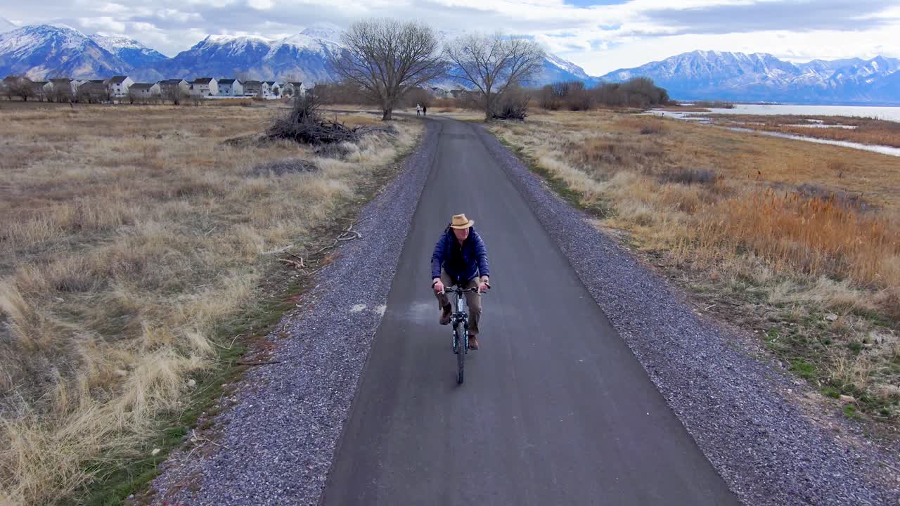 hombre maduro y mayor en bicicleta a lo largo de un sendero natural con pintorescas montañas cubiertas de nieve en la distancia - vista aérea principal