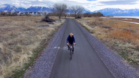 mature, older man bicycling along a nature path with scenic, snowcapped mountains in the distance - leading aerial view