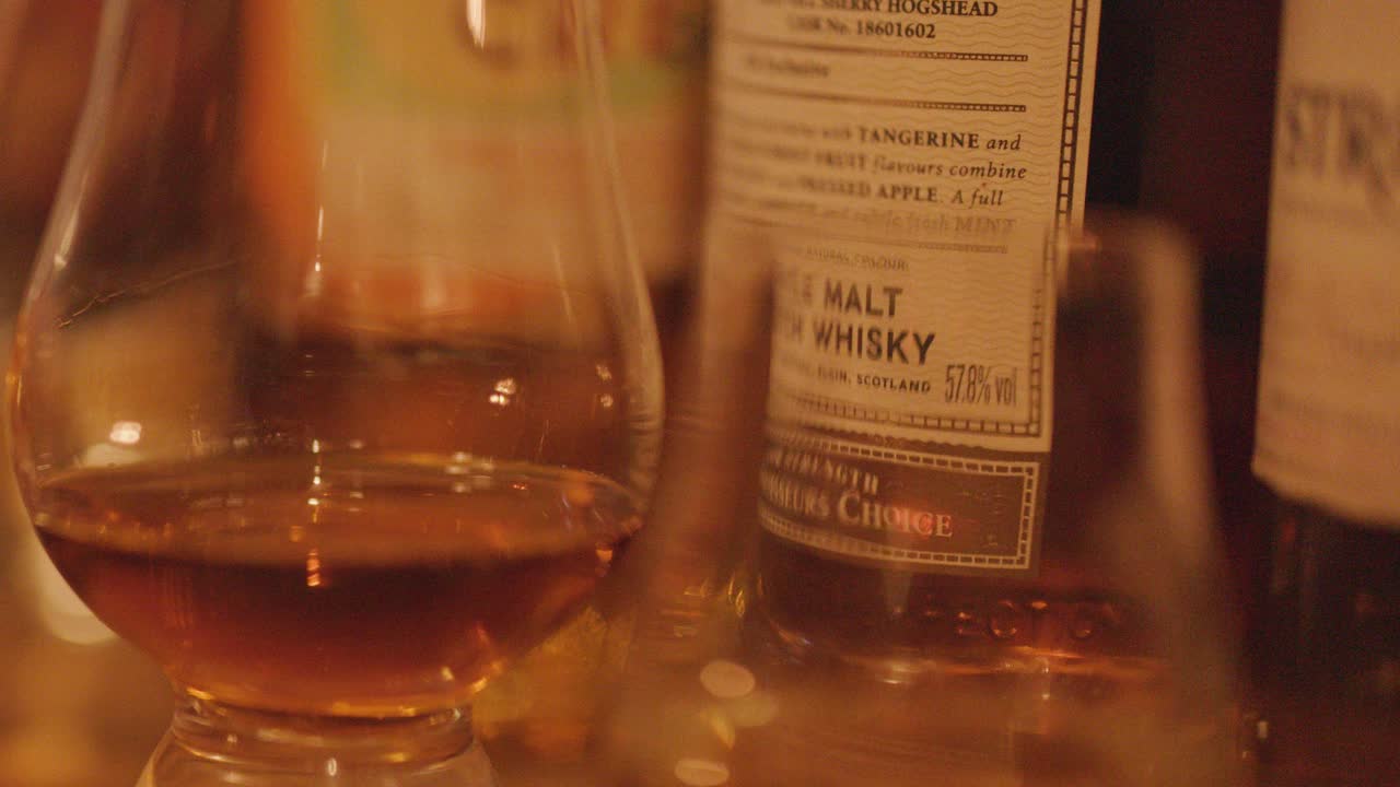 A whisky glass filled with amber spirit stands before several labeled bottles, illuminated by soft, warm lighting in a cozy indoor setting
