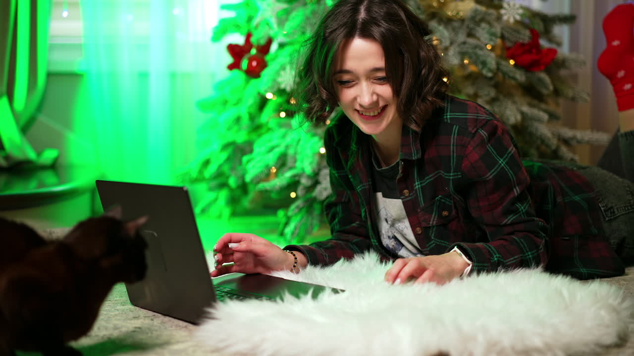 Girl loves Christmas on her laptop. A young woman lies on a cozy white blanket, smiling while using her laptop near a decorated Christmas tree