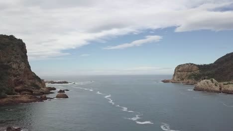 Flight-seaward-through-Knysna-Heads-river-bar-on-a-calm-day,-aerial