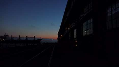 Weitwinkelaufnahme-Eines-Großen-Lagers-Oder-Einer-Fabrik-In-Der-Abenddämmerung-Oder-Bei-Sonnenuntergang,-Während-Ein-Großes-Frachtschiff-In-Der-Ferne-Vorbeifährt