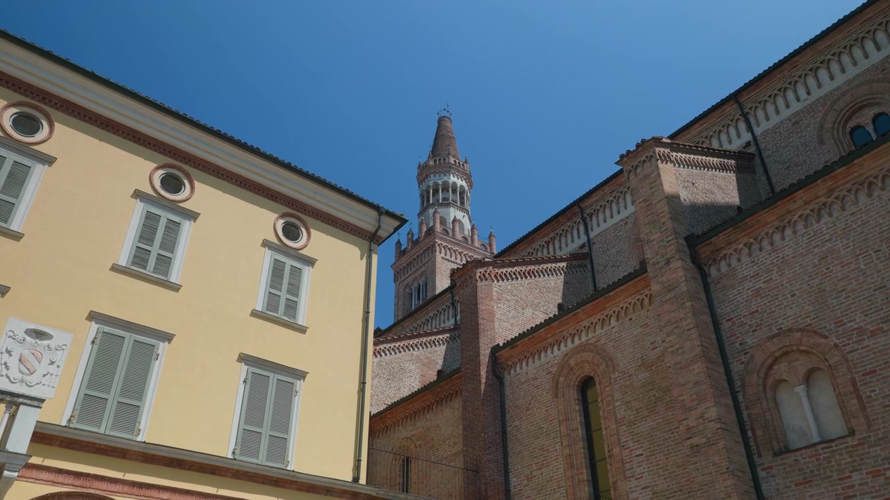 Esterno di cattedrale, campanile e palazzo visti da una piazza storica con una facciata di palazzo gialla e persiane. Architettura medievale italiana in mattoni sotto un cielo azzurro e limpido, panoramica a mano libera