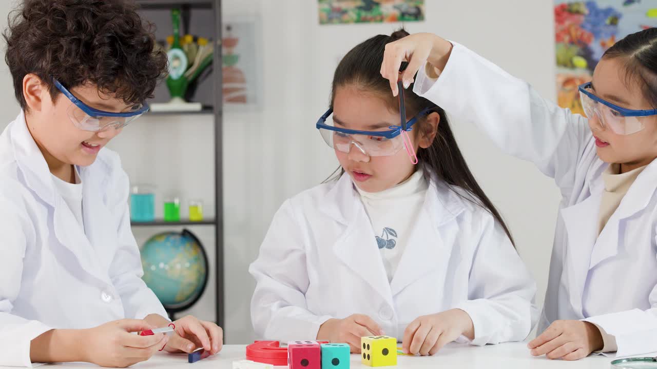 Erilaiset aasialaiset lapset käyttävät laboratoriotakkeja ja suojalaseja tutkiessaan magnetismia luokkahuoneessa.