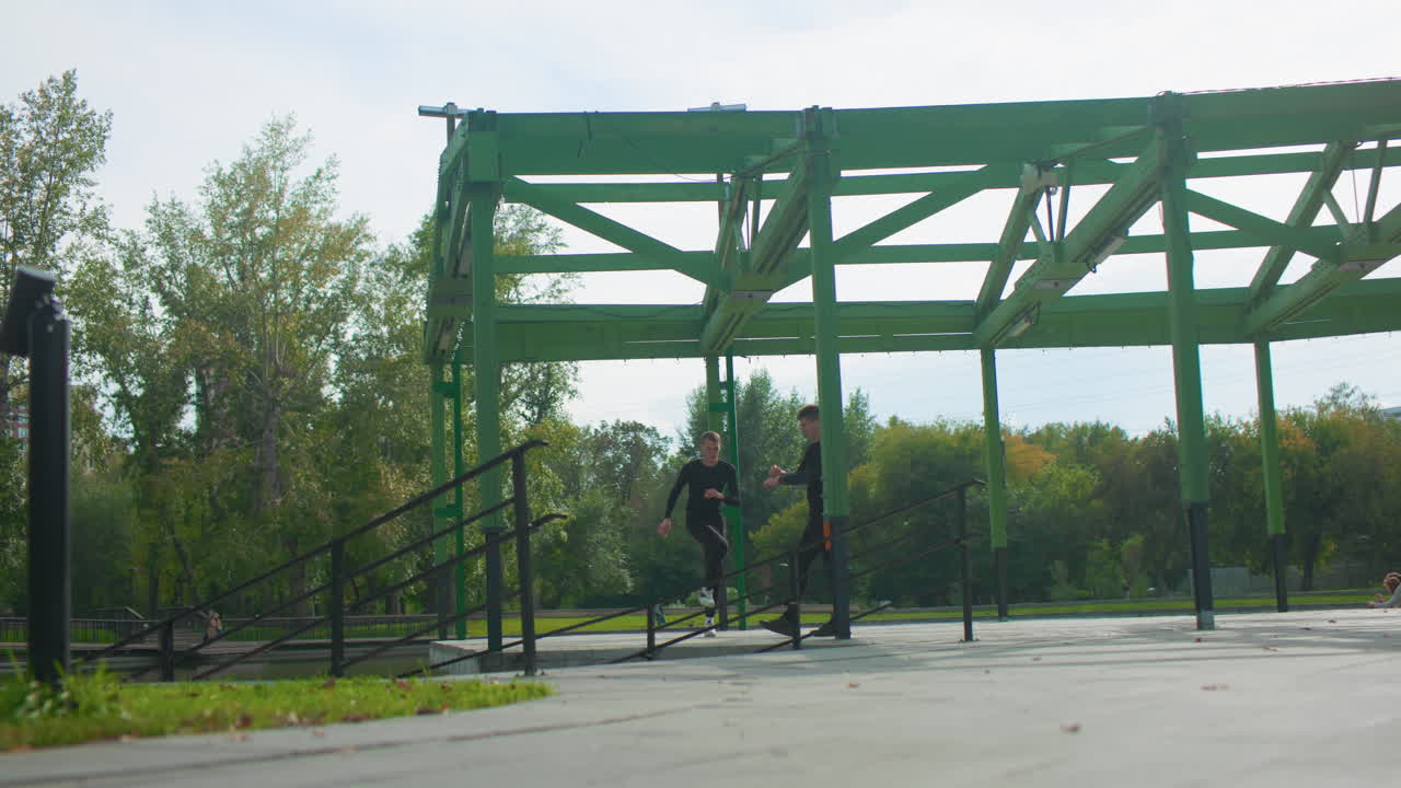 two people training under green pavilion, outdoor fitness meetup on paved path with canopy and trees, stretching and warmup near railing, energetic daytime scene with casual workout vibe