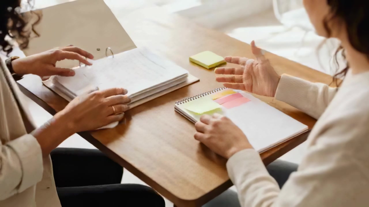 Flipping binder, left woman in blazer handing sheet at wooden table for review with sticky notes
