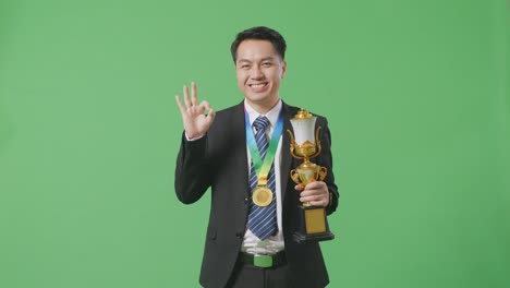 asian business man in a suit and tie with a gold medal and trophy showing okay gesture and smiling to camera as the first winner on green screen background in the studio