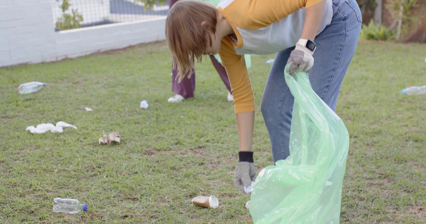 Female volunteers picking up scattered litter on grassy lawn after noticing trash, using green bags