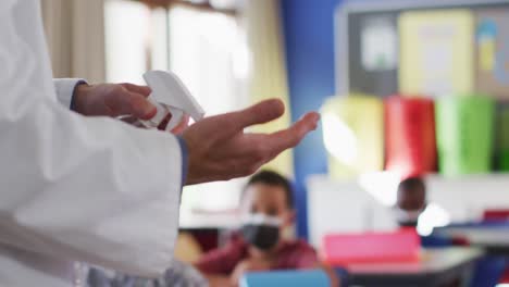 Diverse-teacher-disinfecting-hands-in-classroom,-with-schoolchildren-sitting-wearing-face-masks