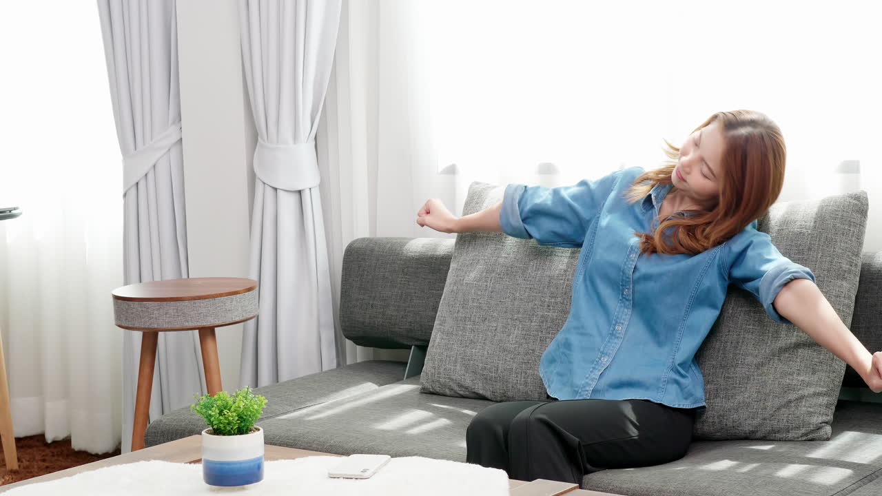 아시아 여성은 퇴근 후 집에 있는 소파에 통증을 완화하기 위해 스마트폰을 탁자 위에 놓고 팔을 쭉 뻗은 채 앉아 있다