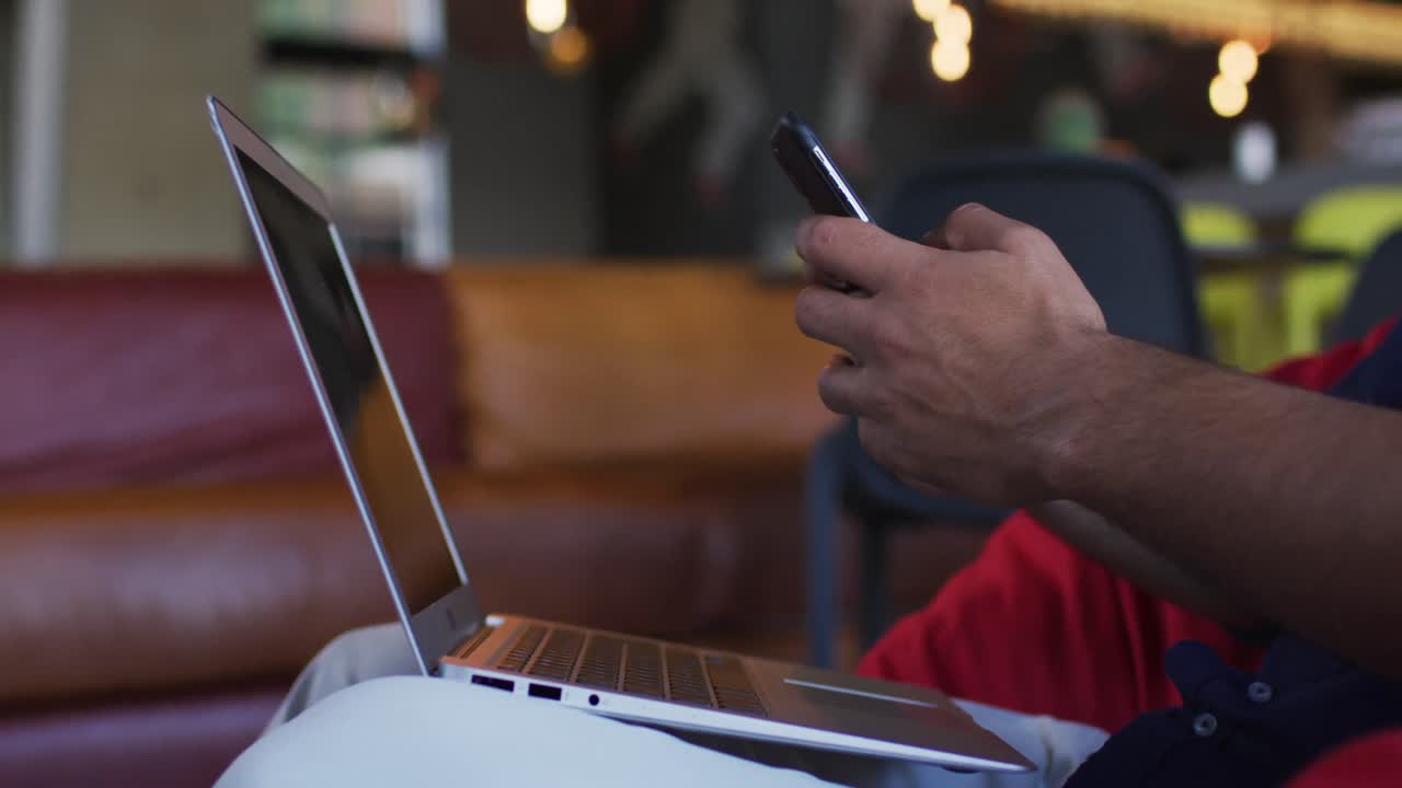 African american man wearing face mask sitting in cafe using smartphone and laptop