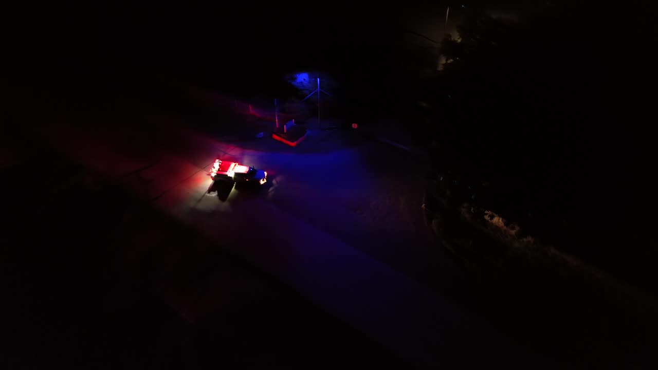 Aerial night view of an emergency vehicle with flashing red and blue lights on a dark road, creating dramatic reflections on the pavement in a quiet, rural setting. Aerial top down shot.