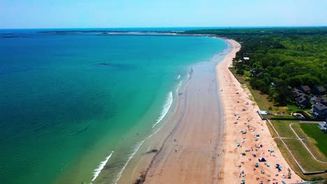 drone aerial of saco, maine coastline with beach crowds. families enjoy waves, kids play in sand near vacation houses. peak-season scene shows beautiful ocean, blue skies, and east coast tourism
