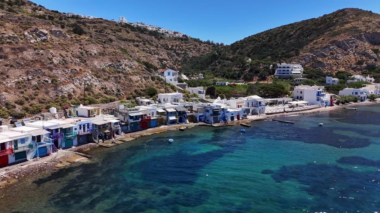 Il villaggio di pescatori di Klima si affaccia su un lungomare roccioso con colorate rimesse per barche syrmata sotto aride colline vulcaniche e limpide acque dell'Egeo, Milos, Grecia, sorvolo obliquo frontale con drone
