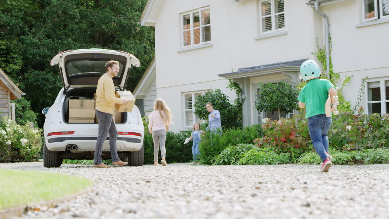 Familie utenfor sitt nye hjem på flyttedagen, laster av esker fra bilen mens sønnen med skateboard løper for å hjelpe.