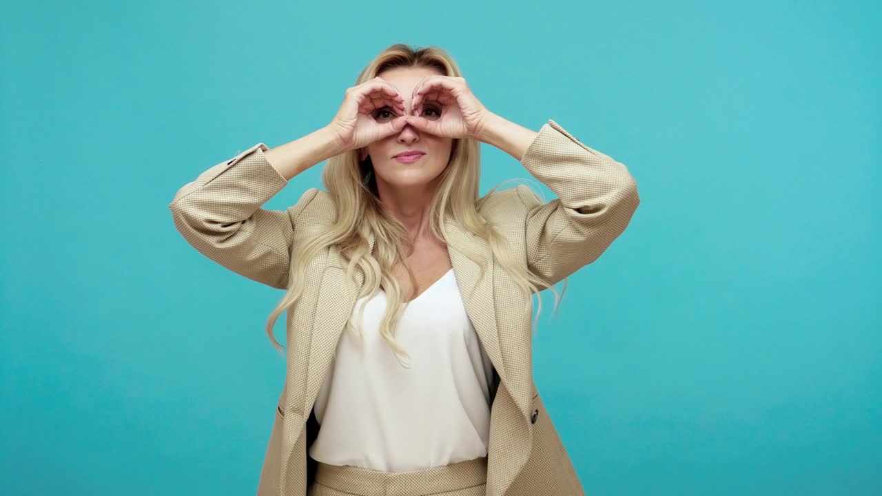mujer curiosa sonriente en chaqueta de negocios sosteniendo las manos cerca de los ojos fingiendo mirar en binoculares, divirtiéndose y preguntándose, espiando