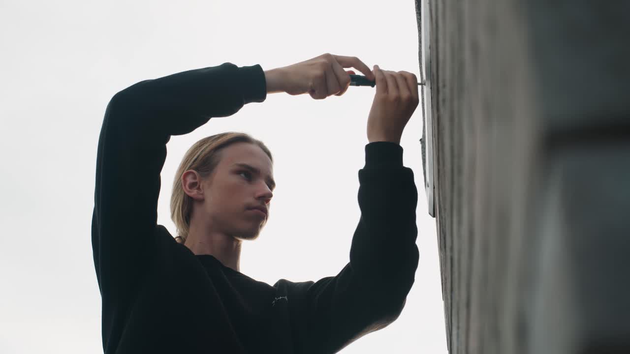 Homem consertando parede de concreto com chave de fenda, close-up de perfil de instalador fixando suporte de metal em fachada urbana sob céu nublado, expressão concentrada, suéter escuro, movimento preciso da mão, telhado.