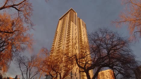 Zoomout-Time-Lapse-De-Nubes-Que-Se-Mueven-Hacia-Arriba-Y-Moderno-Edificio-De-Apartamentos-Futurista-De-Alta-Tecnología-Con-Salón-En-La-Azotea-Que-Brilla-En-Oro,-Puesta-De-Sol-Iluminada-Con-Luz-Natural-Que-Brilla-En-Su-Vista-De-Esquina-Desde-El-Suelo-Hasta-La-Parte-Superior