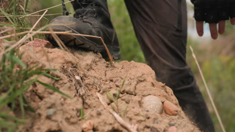 trooper marching on sandy mound focusing on boot plunging into loose sand and grass stems with close up of textured soil stones and gloved hand grip hinting at military tension in wilderness terrain