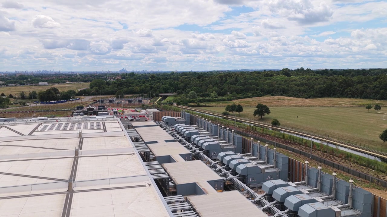 Store industrielle køleenheder langs taget af en datacenterbygning beliggende nær landlige marker og træer i klart dagslys med en synlig fjern horisont under blå himmel