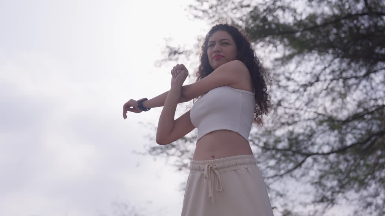 A slow-motion low-angle view of a South Asian young woman in a white crop tank top doing workout exercises under a cloudy sky with tree branches