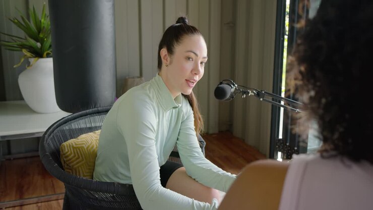 Two women in studio on guest cue, host turning, showing tablet screen, gesturing with boom mic