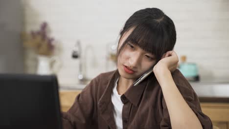 closeup view of a chinese female holding mobile phone between ear and shoulder and looking at computer screen while discussing work with colleague at home