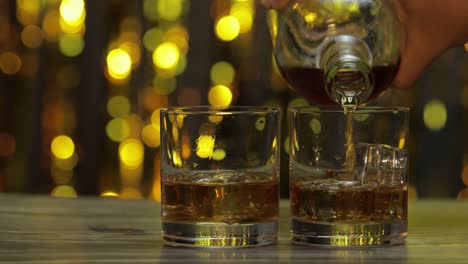 pouring of golden whiskey, cognac or brandy from bottle into glass with ice cubes. shiny background