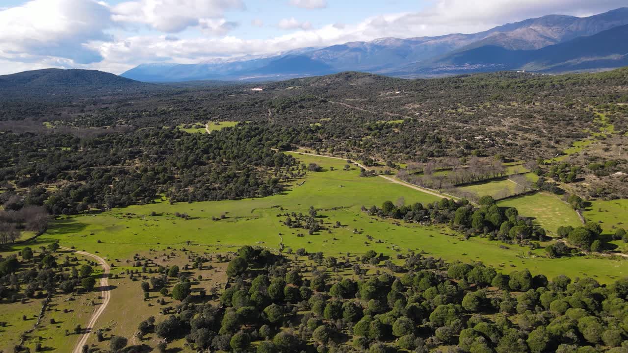vuelo en un valle en invierno con montañas en el fondo y un cielo azul con nubes viendo prados verdes con carreteras y bosques de pinos y otra diversidad de árboles, algunos sin hojas españa