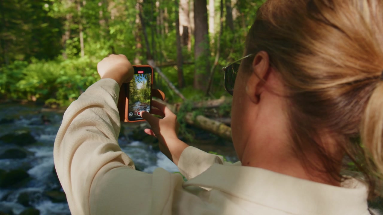 Kvinde filmer skovbæk med telefon. Hun fokuserer intenst og nyder naturens skønhed. Et overhead-billede viser hende i solbeskinnet skov. Ideel til rejse-, eventyr- eller økoturisme-indhold