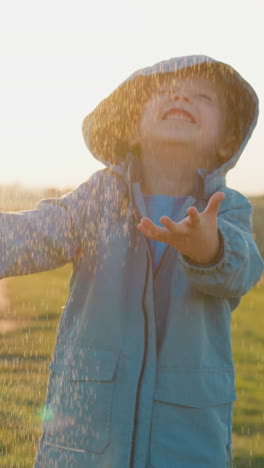 kid holds palms under warm spring rain in field. cheerful toddler boy wearing raincoat has fun at pouring rain at countryside. stormy weather on river bank