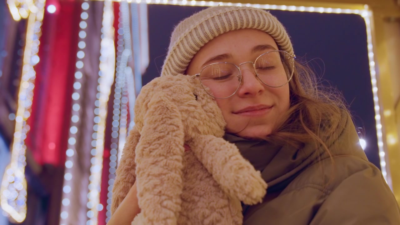 Jovem mulher abraçando um coelho de pelúcia sob as luzes cintilantes do mercado, usando gorro e parka, fechando os olhos em contentamento pacífico, um retrato íntimo de noite de inverno com brilho quente e clima nostálgico.
