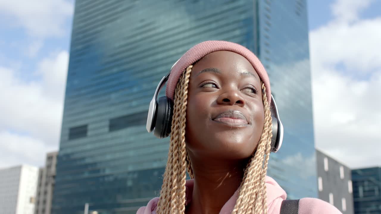 En kvinde i lyserød hættetrøje lytter til musik i hovedtelefoner, vender sig og smiler mod et glastårn, med plads til tekst.