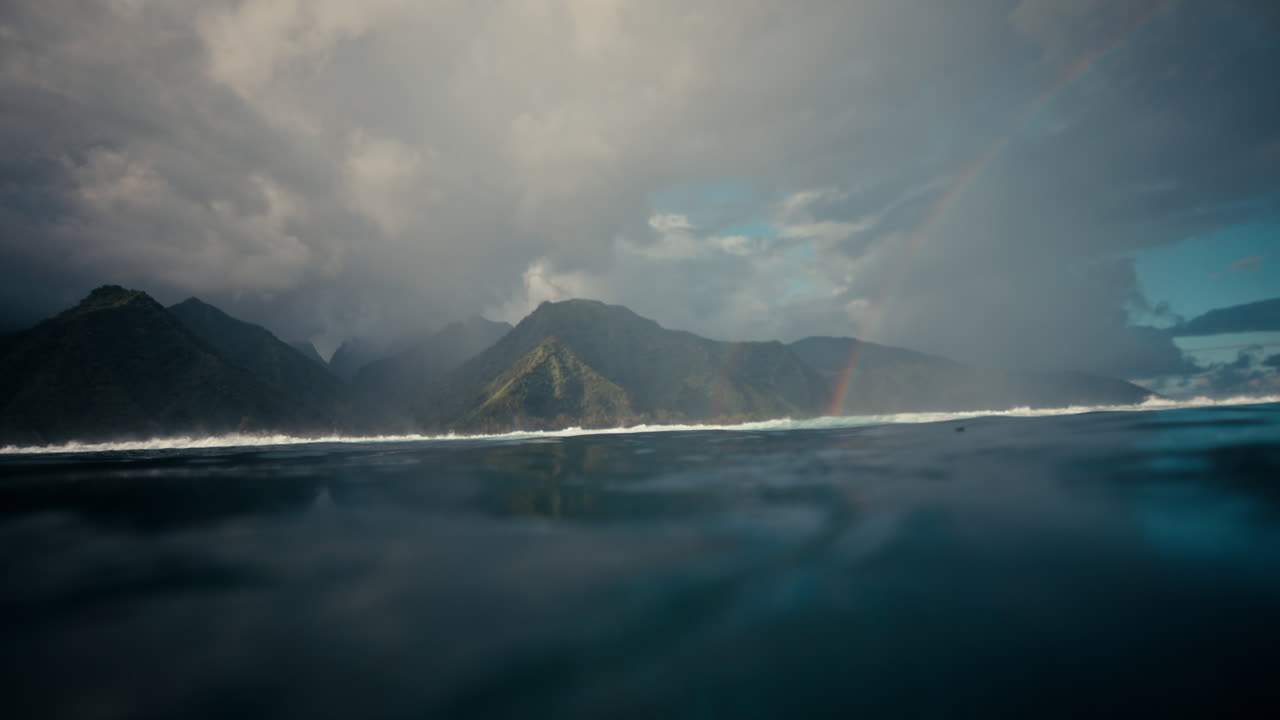Cảnh mặt biển nhìn về phía những ngọn núi nhiệt đới xanh tươi ở Tahiti dưới những đám mây ấn tượng và cầu vồng xa xăm.