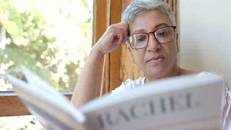 Feliz-Mujer-Birracial-Mayor-En-El-Sofá-Leyendo-Un-Libro-En-La-Ventana-De-Casa,-Cámara-Lenta