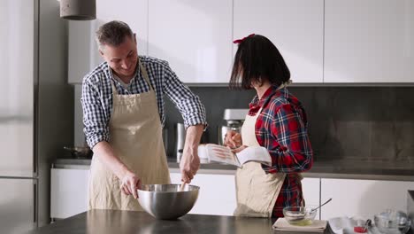 Un-Joven-Preparando-Comida-Y-Una-Mujer-Mostrándole-Algo-En-Un-Libro-De-Recetas.