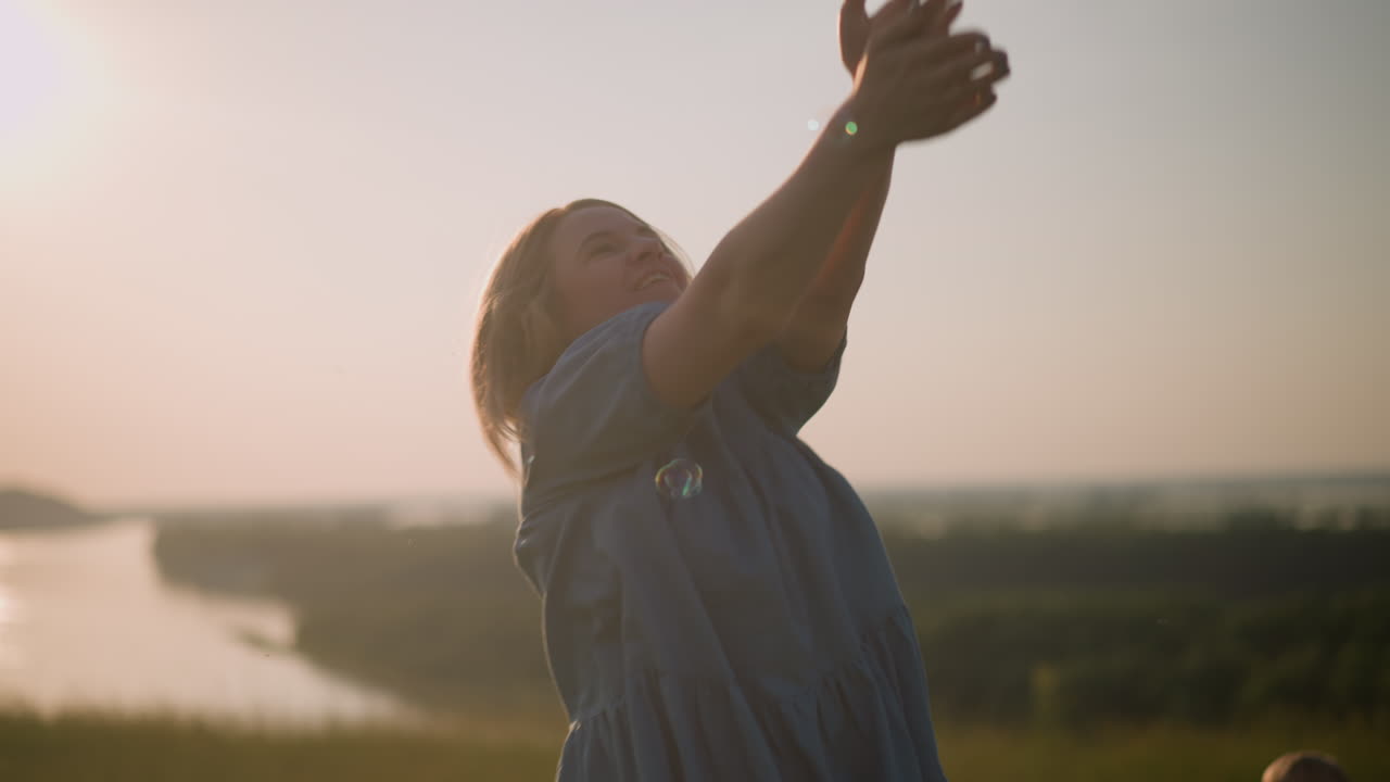 A woman in a blue dress jumps gracefully in a sunset field, reaching out with joy to touch floating bubbles in the warm evening light, capturing a serene moment of playful happiness in nature