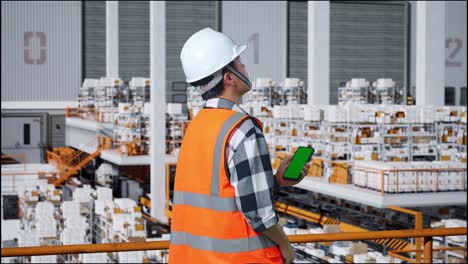 back view of asian male engineer with safety helmet standing in the warehouse with shelves full of delivery goods. zoom in green screen smartphone and looking around the storage