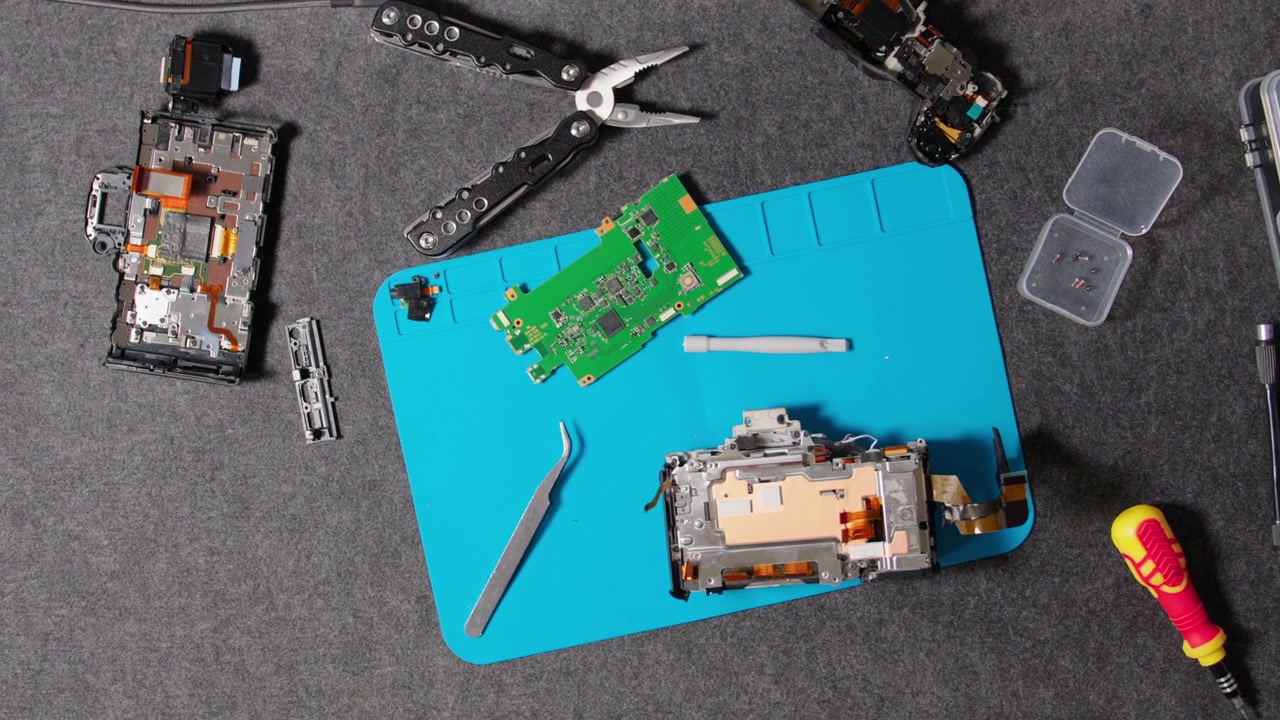 Disassembled camera body and circuit board with various specialized tools for electronics repair and maintenance on a gray mat.
