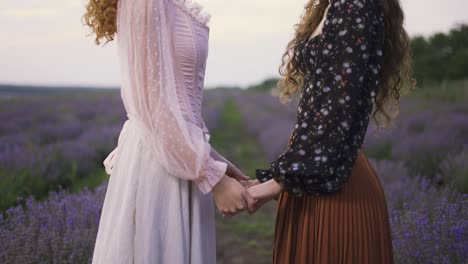 Two-gorgeous-women-are-standing-among-lavender-field,-holding-hands-and-looking-to-each-other