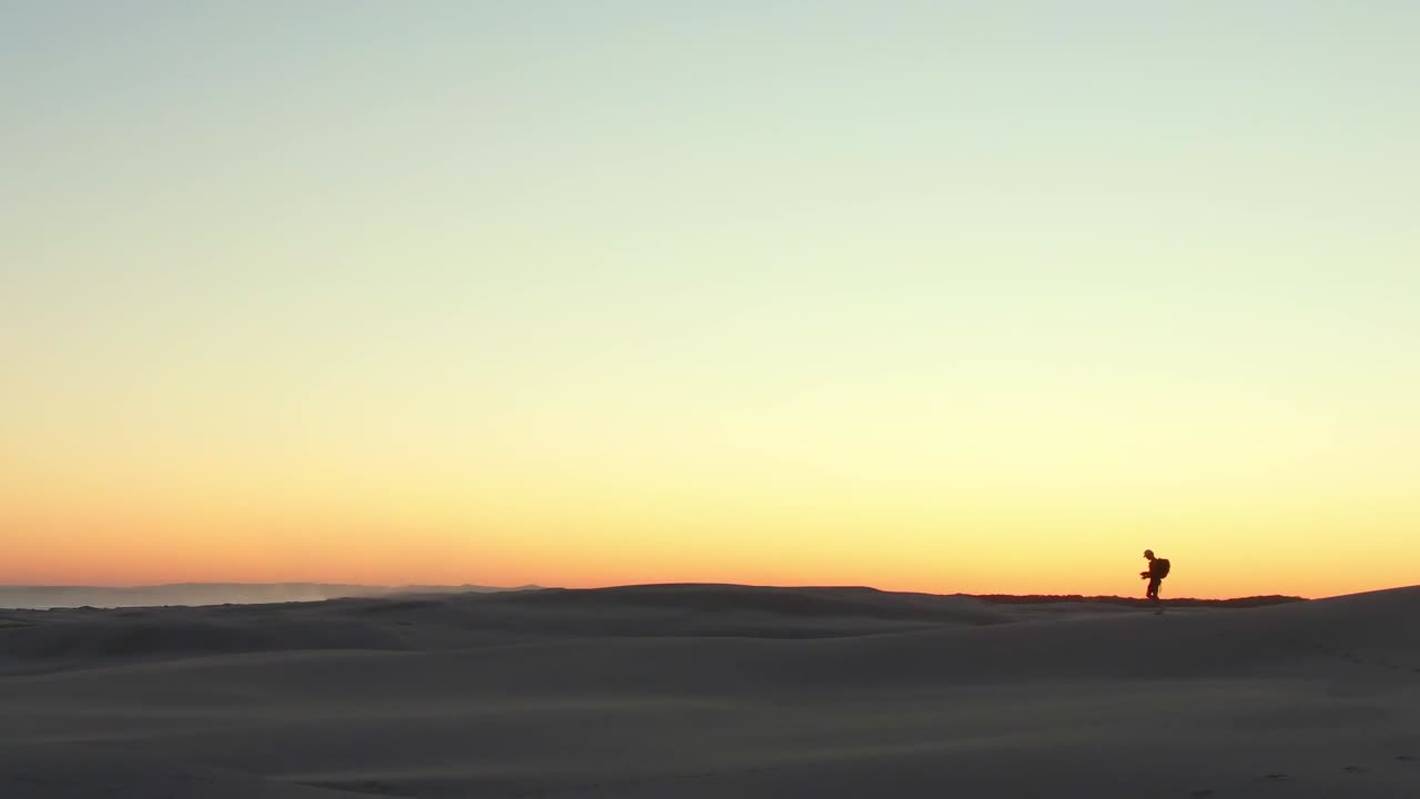 A man hikes along the top of a sand dune with the sunset behind him castings a silhouette against the golden orange sky.