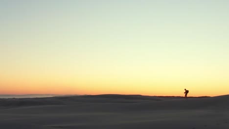 a man hikes along the top of a sand dune with the sunset behind him castings a silhouette against the golden orange sky.