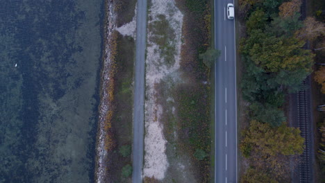 overhead shot of a two-lane road with vehicles between a sandy area and a forest with autumn foliage
