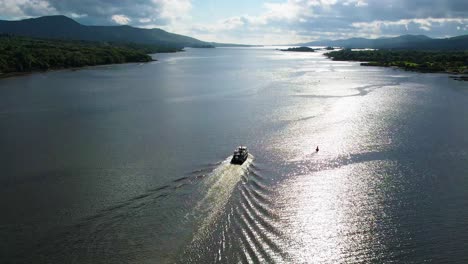 Eine-Aufsteigende-4K-Luftaufnahme-Eines-Touristenbootes,-Das-Im-Sonnenschein-In-Der-Bucht-Von-Kenmare-Auf-Das-Offene-Meer-Zusteuert