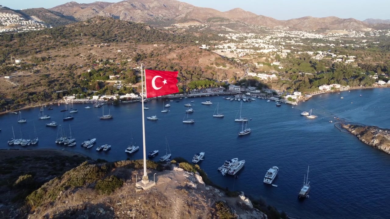 Luftfoto baglæns af tyrkisk flag, der blafrer, med Gümüşlük i baggrunden.