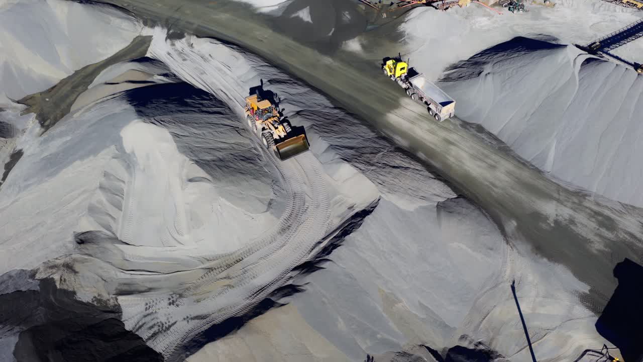 Dump truck driving between gravel piles as front loader backs up on aggregate, pullback aerial reveal