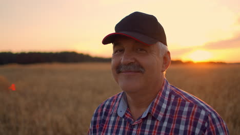 Retrato-De-Un-Granjero-Adulto-Mayor-En-Un-Campo-De-Grano-Mirando-La-Cámara-Y-Sonriendo-Al-Atardecer.-El-Conductor-Del-Tractor-Se-Quita-La-Gorra-Y-Mira-A-La-Cámara-En-Cámara-Lenta