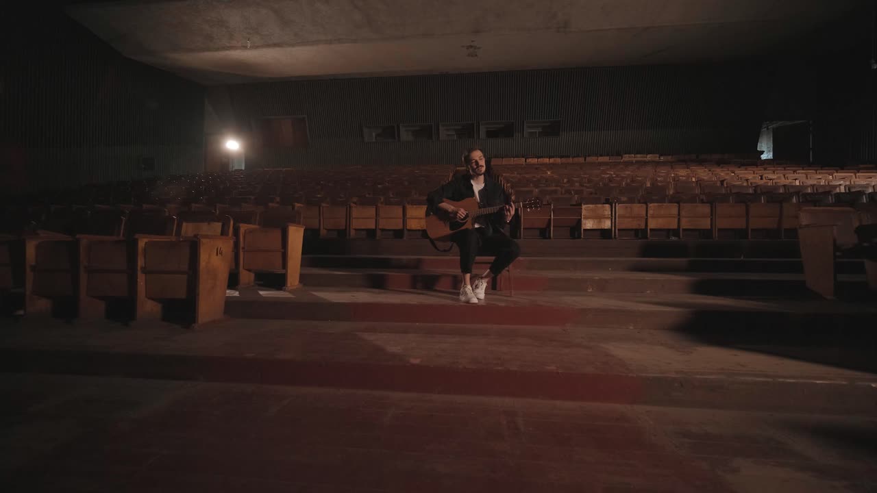 un chico guapo toca una guitarra acústica en un cine abandonado. el músico canta una canción y acompaña en la guitarra